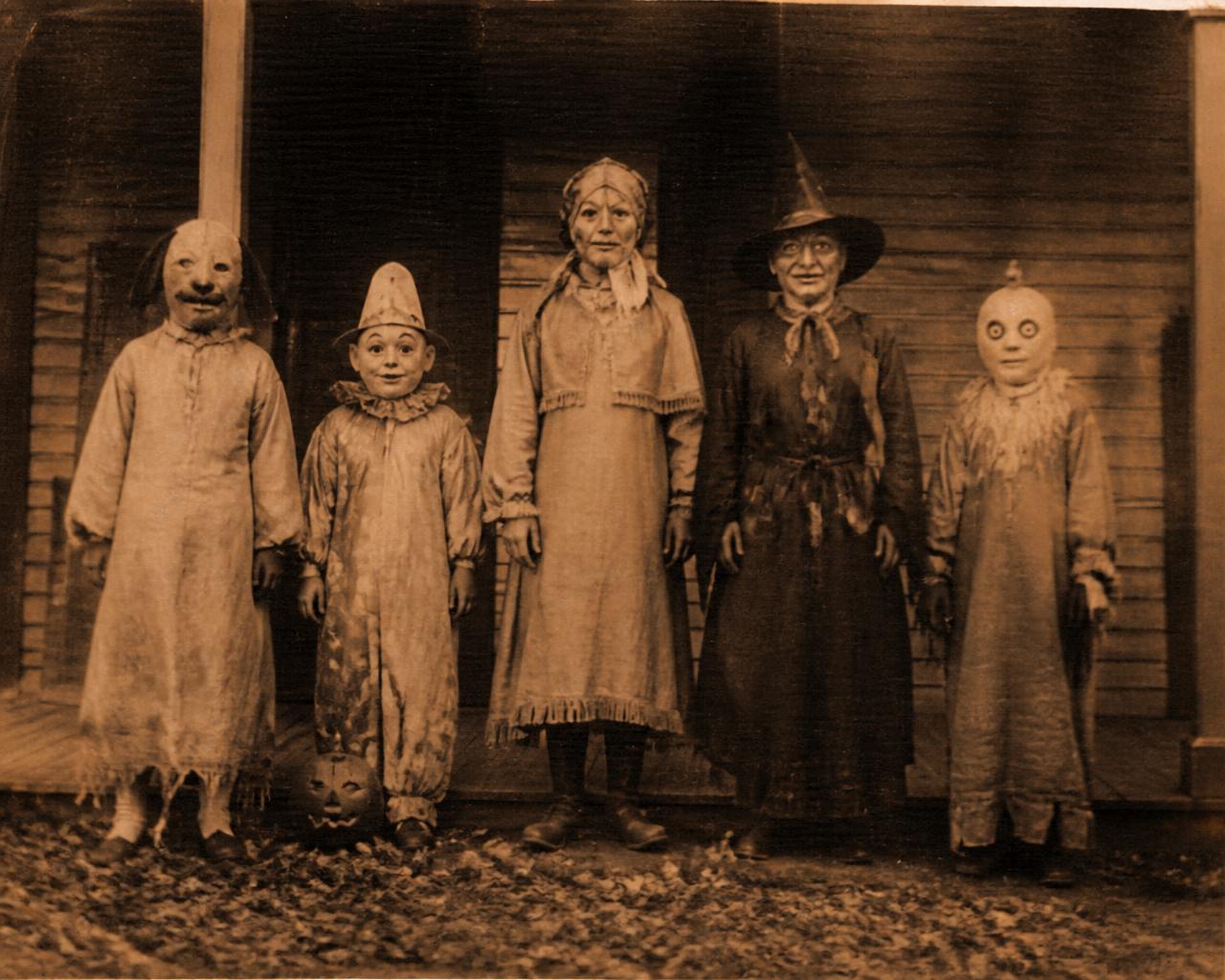 Sepia-toned vintage photograph of five figures in homemade cloth Halloween masks standing on a dim farmhouse porch